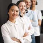 Smiling diverse customer service team with headsets standing in a line indoors.
